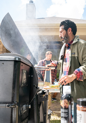 Person tending a large smoker grill at an outdoor gathering. Smoke rises from the grill while food cooks inside. Other people and picnic-style activity are visible in the background.