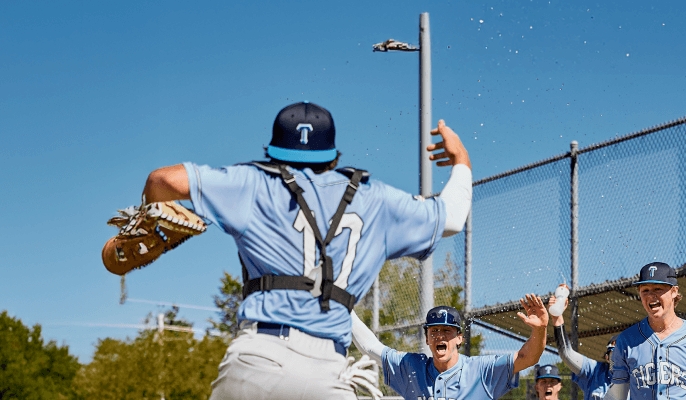 Group of baseball players in light blue 'TIGERS' jerseys celebrating near a field fence, with one player in catcher’s gear and others raising hands for high-fives.