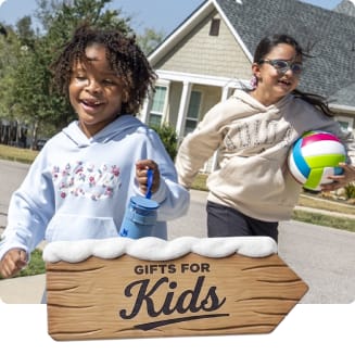Two children running outdoors in a neighborhood, one carrying a colorful volleyball and the other holding a water bottle. A wooden holiday sign with snow reads “Gifts for the Kids.”