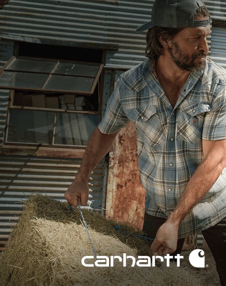 Person wearing a plaid short‑sleeve plaid Cahartt work shirt handling hay outside a corrugated metal barn, with Carhartt logo displayed on bottom right of image.