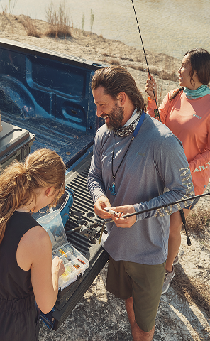 Three people gathered around the open tailgate of a pickup truck outdoors. One person stands beside the truck bed while two others look toward the open storage area. Dry grass and fencing are visible in the background.