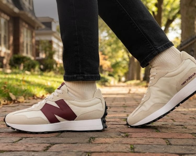 Cream and burgundy New Balance sneaker worn with black pants on a brick walkway, with New Balance logo.
