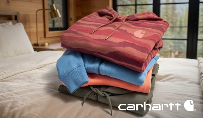 Stack of folded Carhartt hooded sweatshirts in multiple colors displayed on a bed.