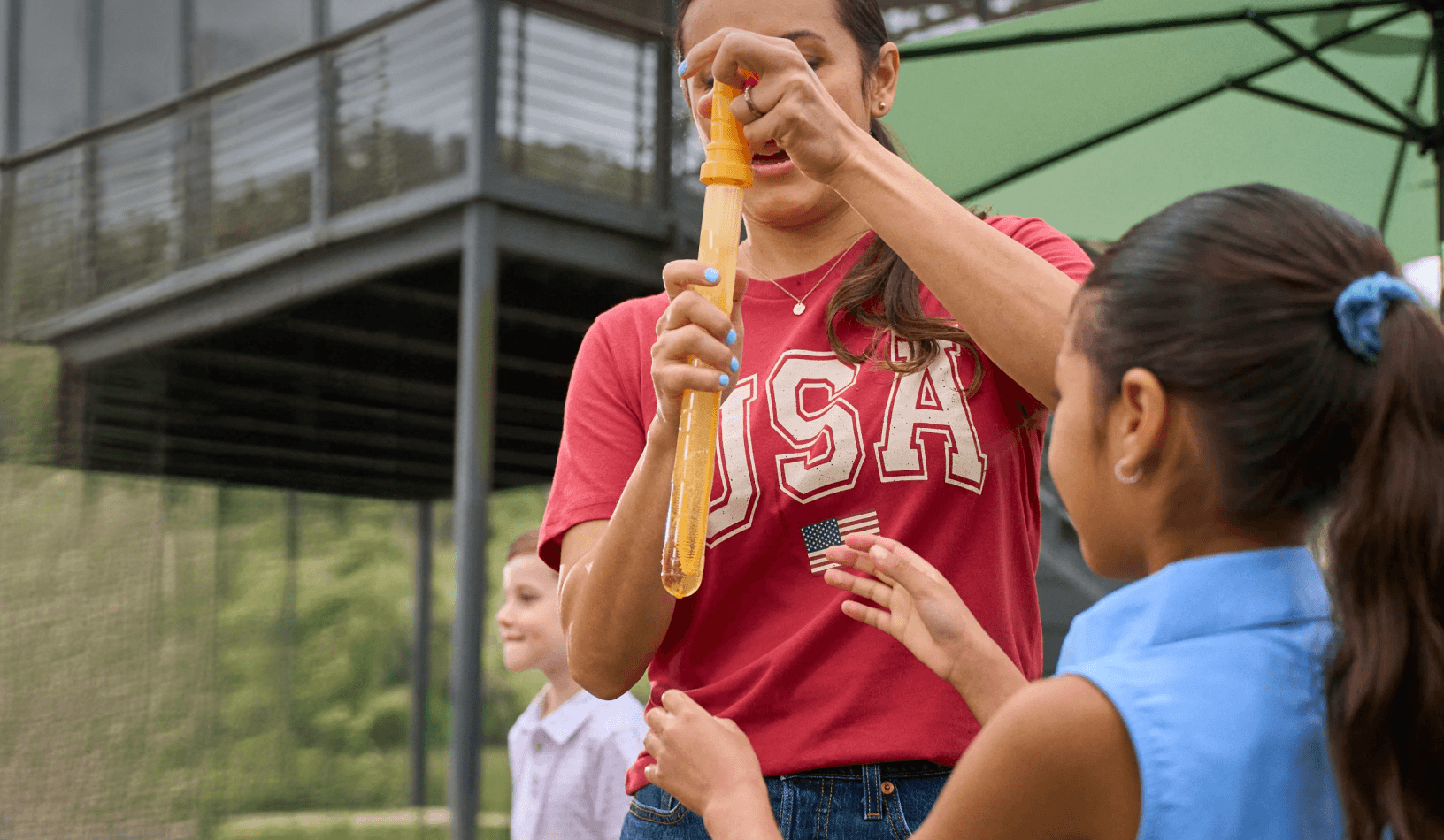 People outdoors, with one person wearing a red USA shirt and holding a bubble wand near a child.