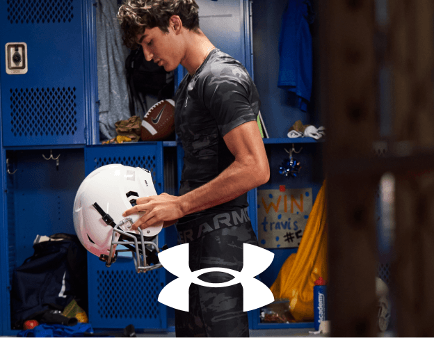 Person holding a basketball in a gym while standing beside sports equipment and Under Armour branding.