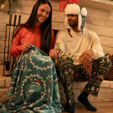 Two people sitting by a fireplace wearing Magellan Outdoors holiday apparel and holding a patterned blanket.