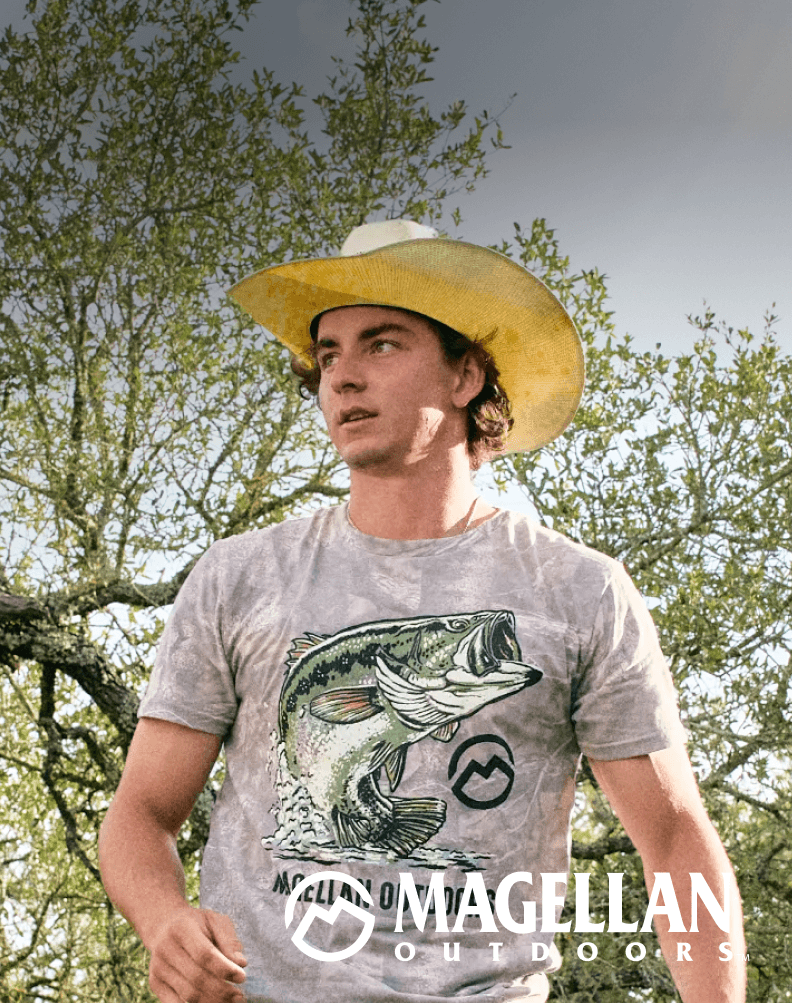 Person wearing a washed camo graphic T-shirt and wide brim hat standing outdoors among trees.
