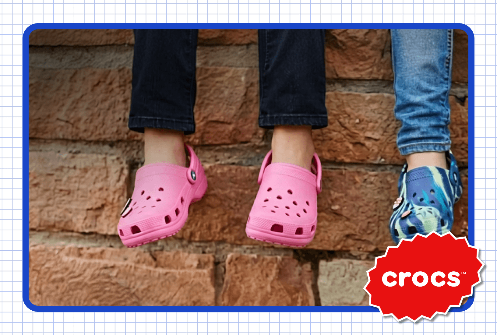Two pairs of feet, one wearing pink and one wearing blue Crocs shoes sitting on a brick wall.