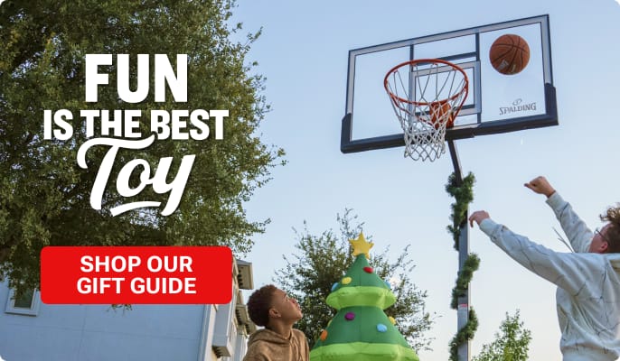 People playing basketball outdoors beneath a hoop decorated with holiday garland beside a large inflatable Christmas tree with overlaid text: Fun is the Best Toy and button: Shop our gift guide.