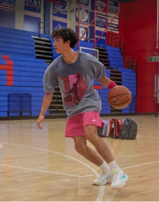 Person dribbling a basketball on an indoor gym court with bleachers in the background.