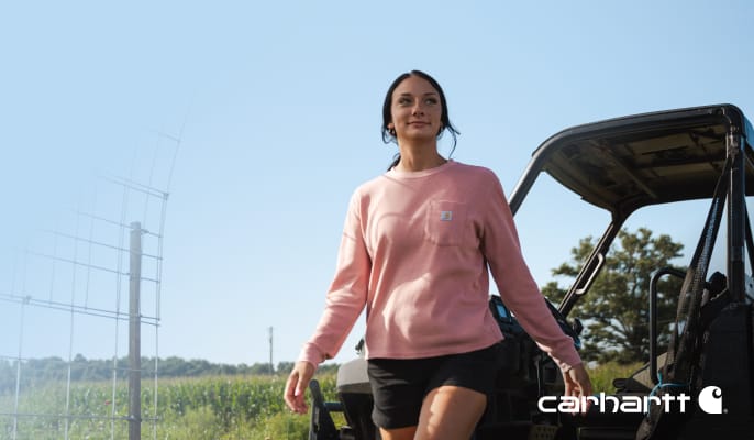 Farmer Grayce Emmick walking outdoors beside a utility vehicle in a pink Carhartt sweatshirt and black shorts.