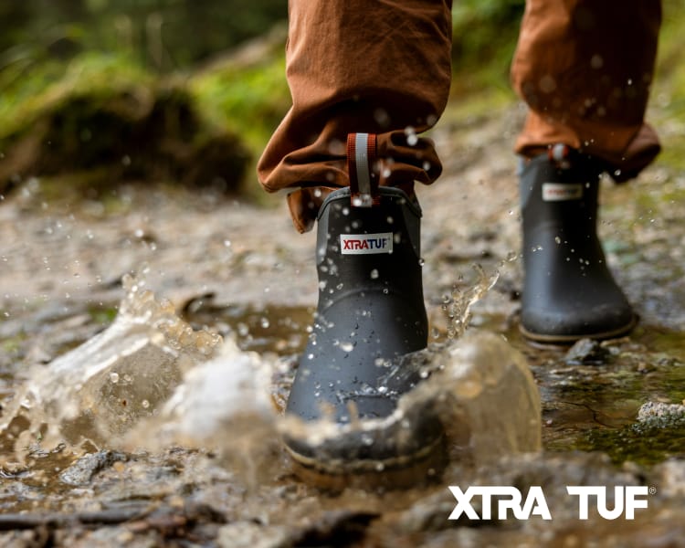 Close-up of waterproof XTRATUF deck boots splashing through a shallow stream with water droplets in motion. XTRATUF logo appears in the lower-right corner.