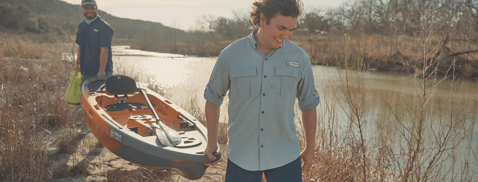 Person pulling a kayak along the edge of a lake with another person walking nearby while wearing Magellan Outdoors Laguna Madre shirts.
