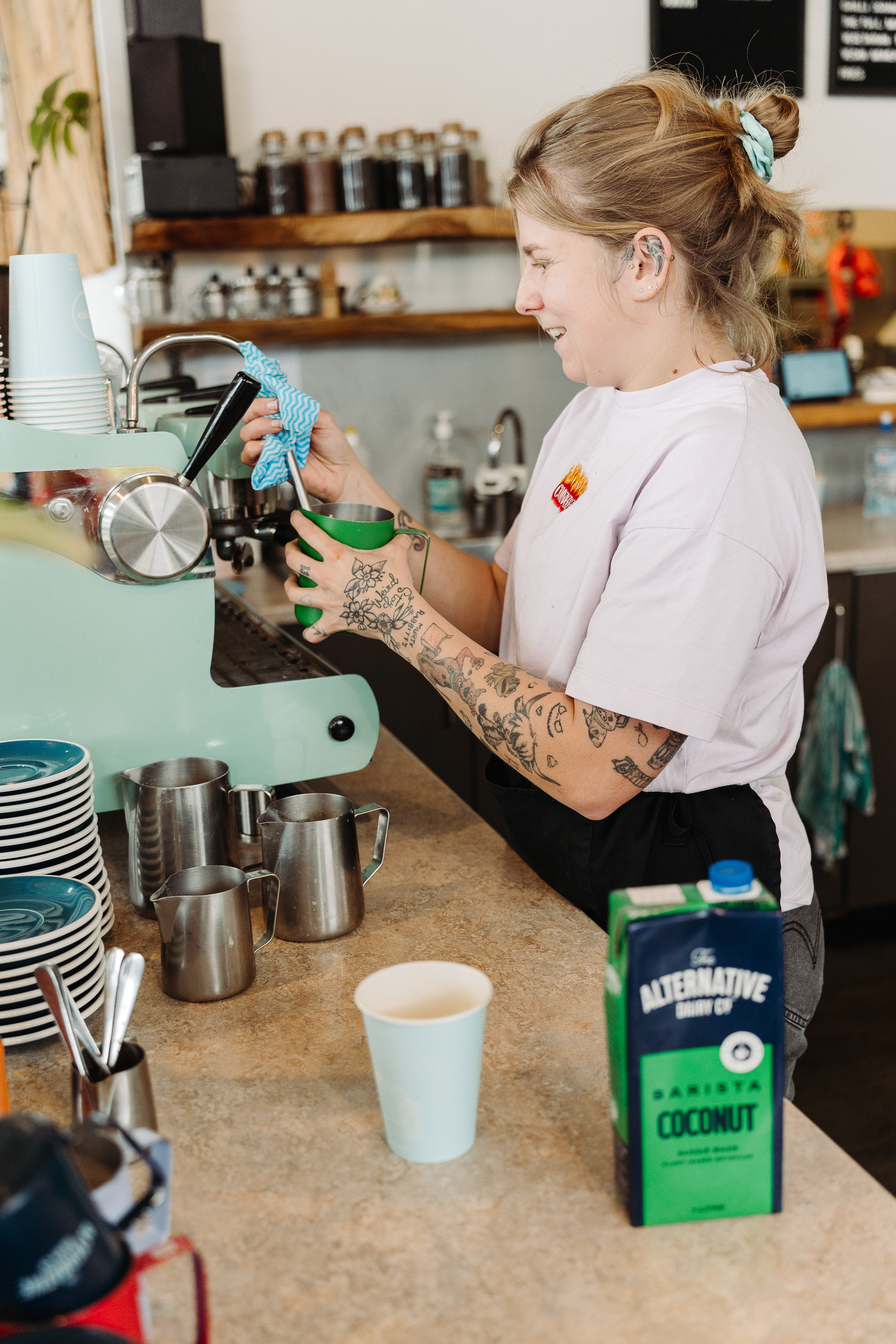 A barista wipes down an espresso machine while holding a green milk frothing pitcher behind a café counter, with cups, jugs, and coffee supplies in the foreground.