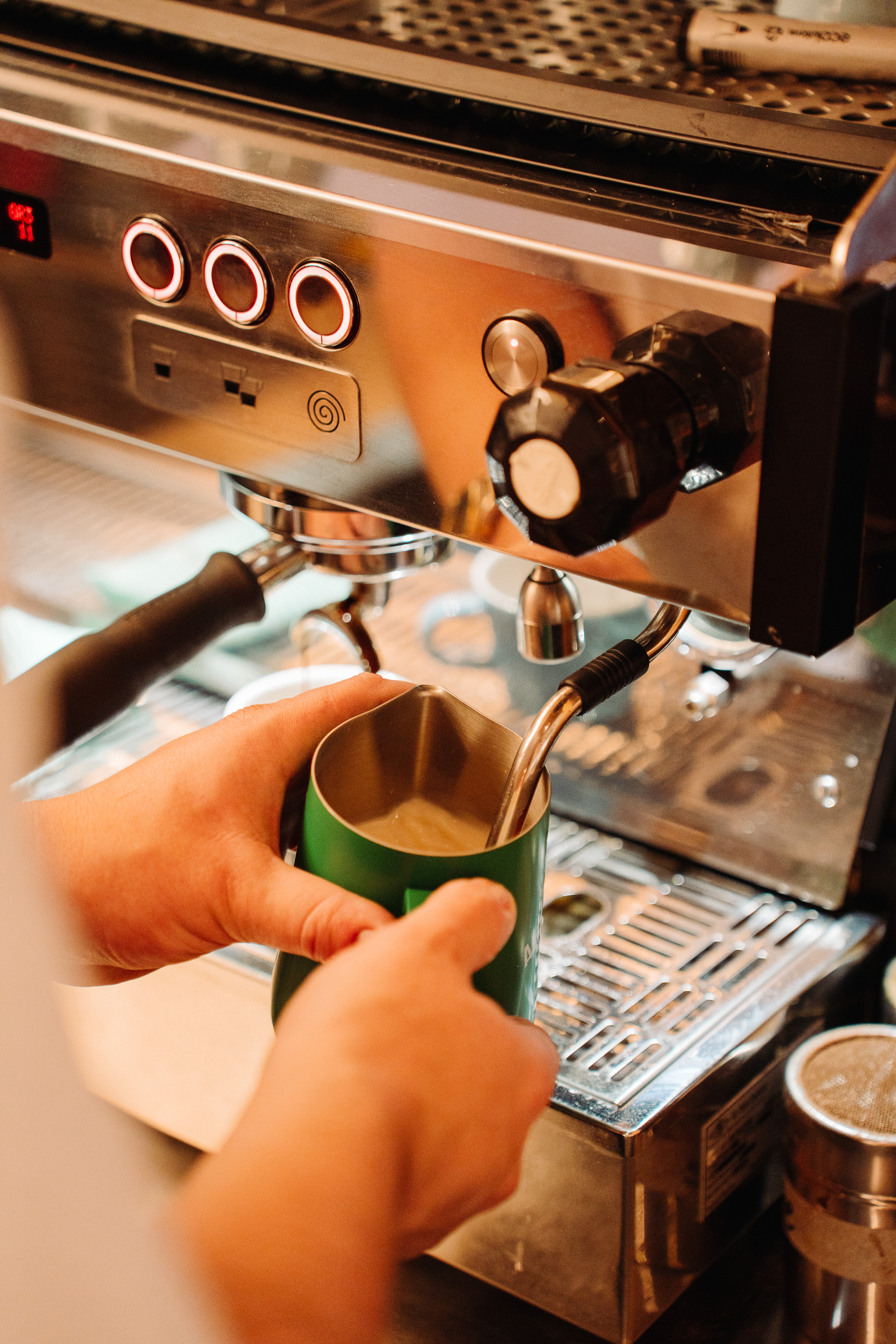 Close-up of an espresso machine steaming milk into a green metal pitcher, with a barista’s hands holding the jug under the steam wand.