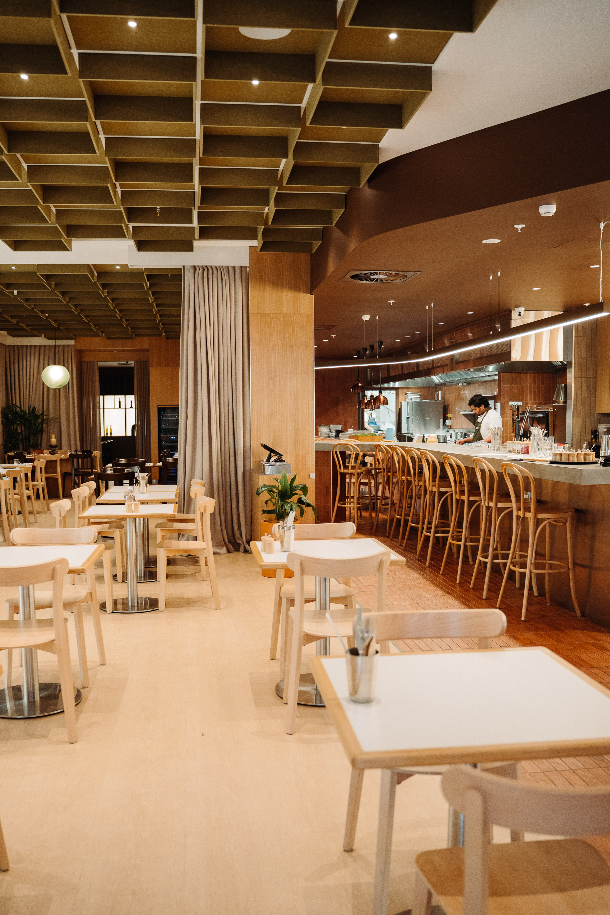 Warm, modern restaurant interior with an open kitchen and long bar lined with wooden stools, set beneath a geometric ceiling feature and soft ambient lighting.