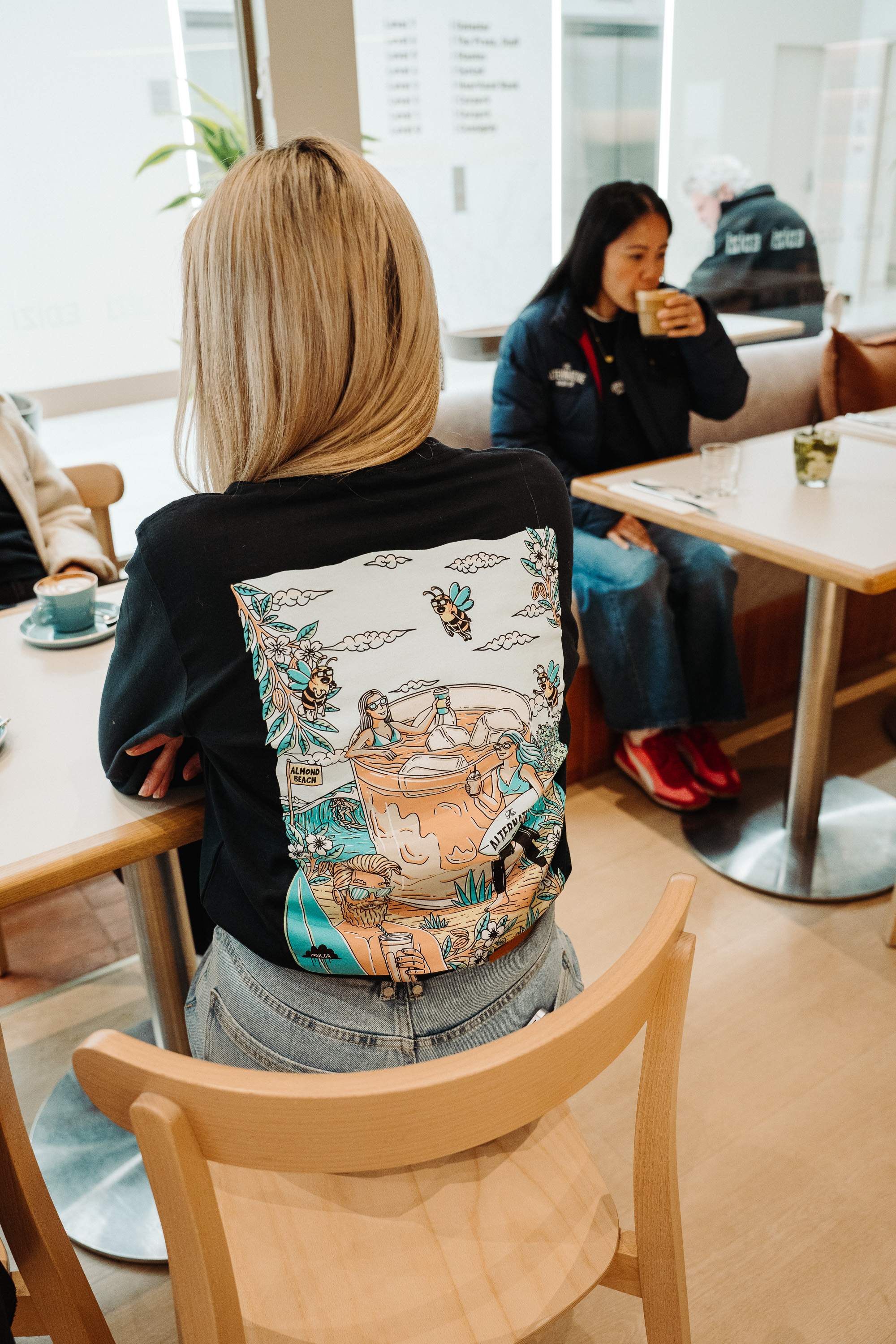 A candid indoor café scene with two people seated at light wooden tables; one person is seen from behind wearing a black shirt with a colorful illustrated graphic on the back, while another person in the background sips a hot drink.
