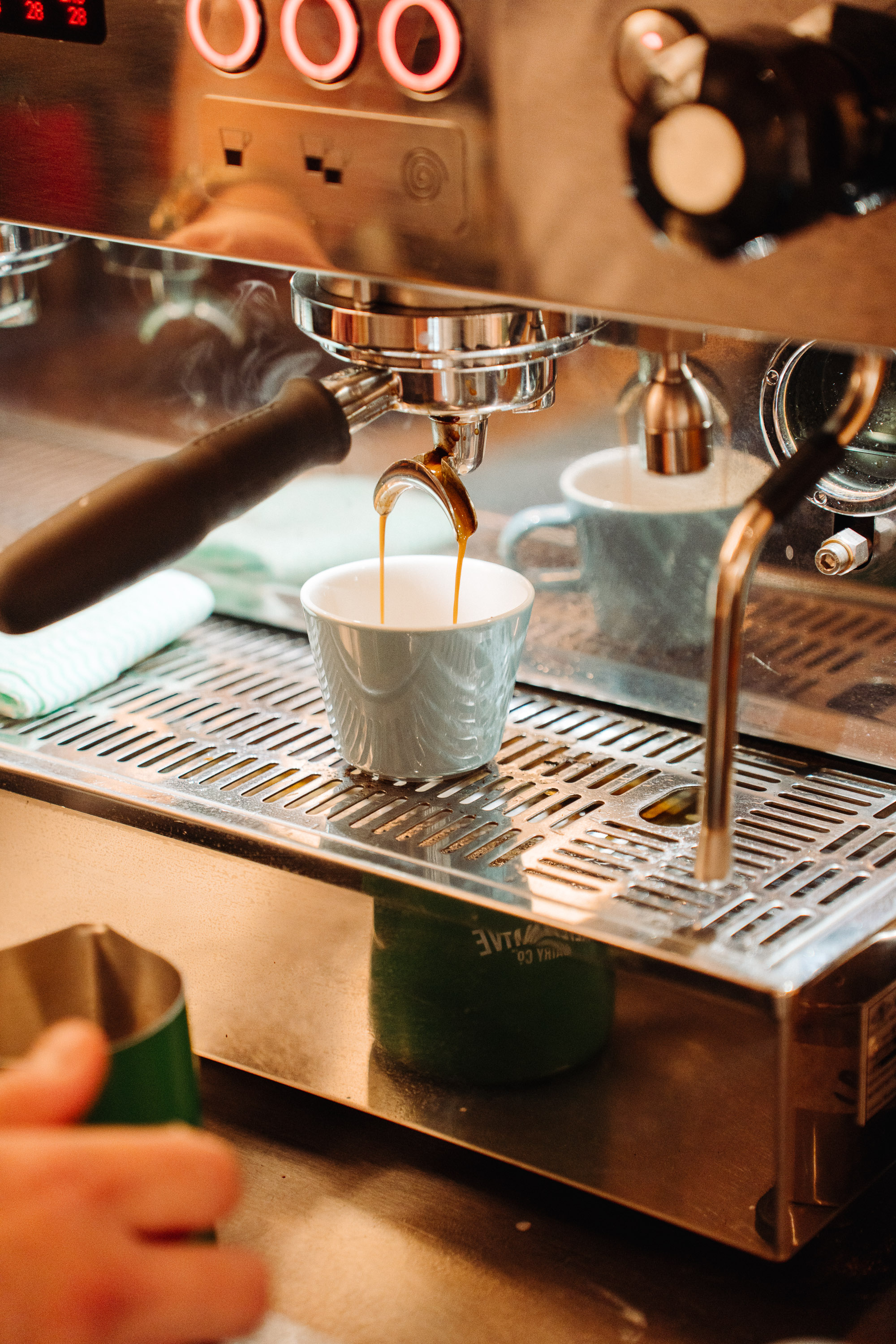Close-up of an espresso machine pulling a shot, with two thin streams of coffee flowing from the portafilter into a ceramic cup on the drip tray. Warm cafe lighting reflects off the stainless-steel surfaces and steam wand nearby.