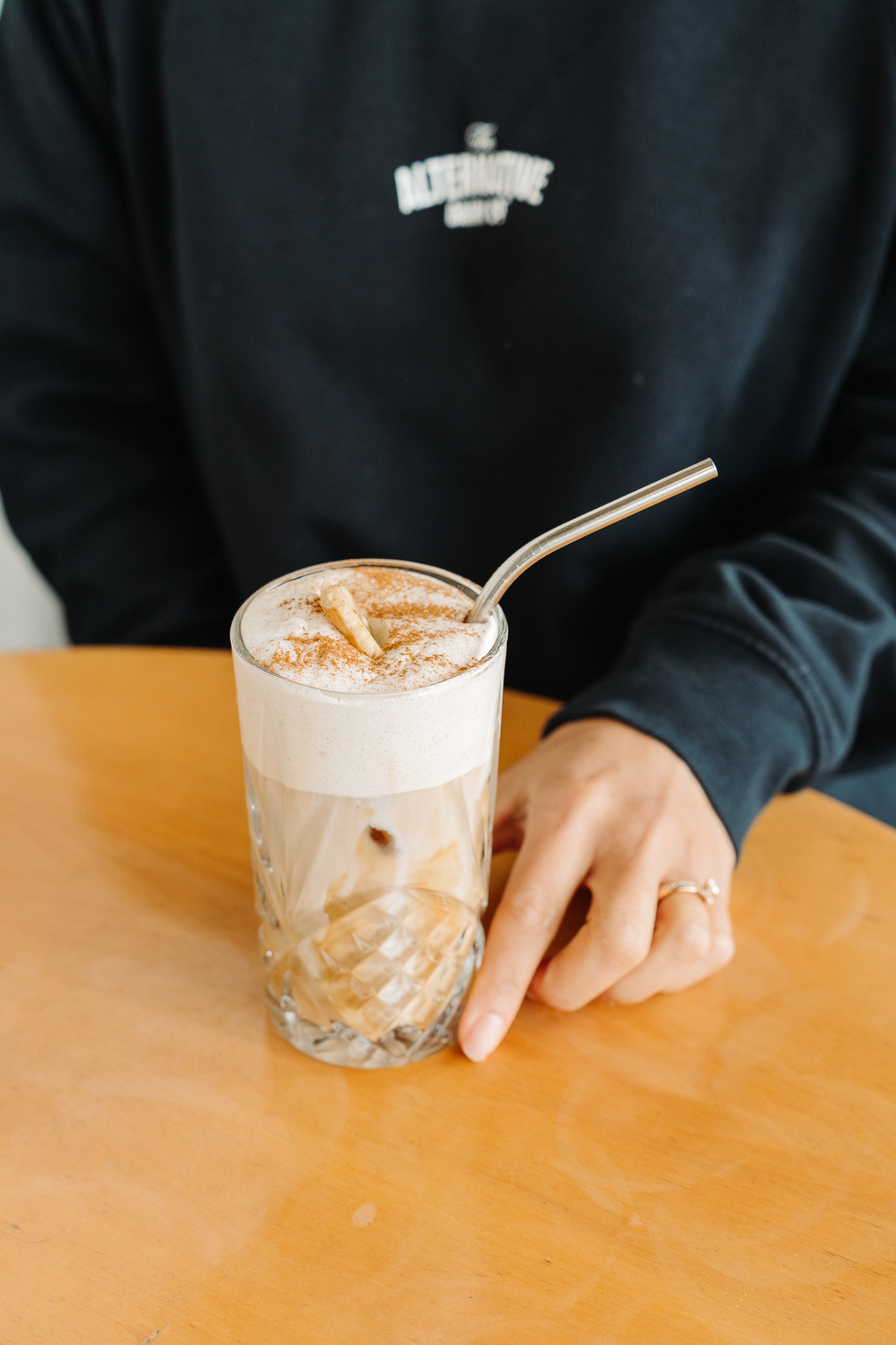 Close-up of an iced latte in a glass with a thick layer of foam and a metal straw, sitting on a wooden table as someone in a dark sweatshirt rests a hand beside it.