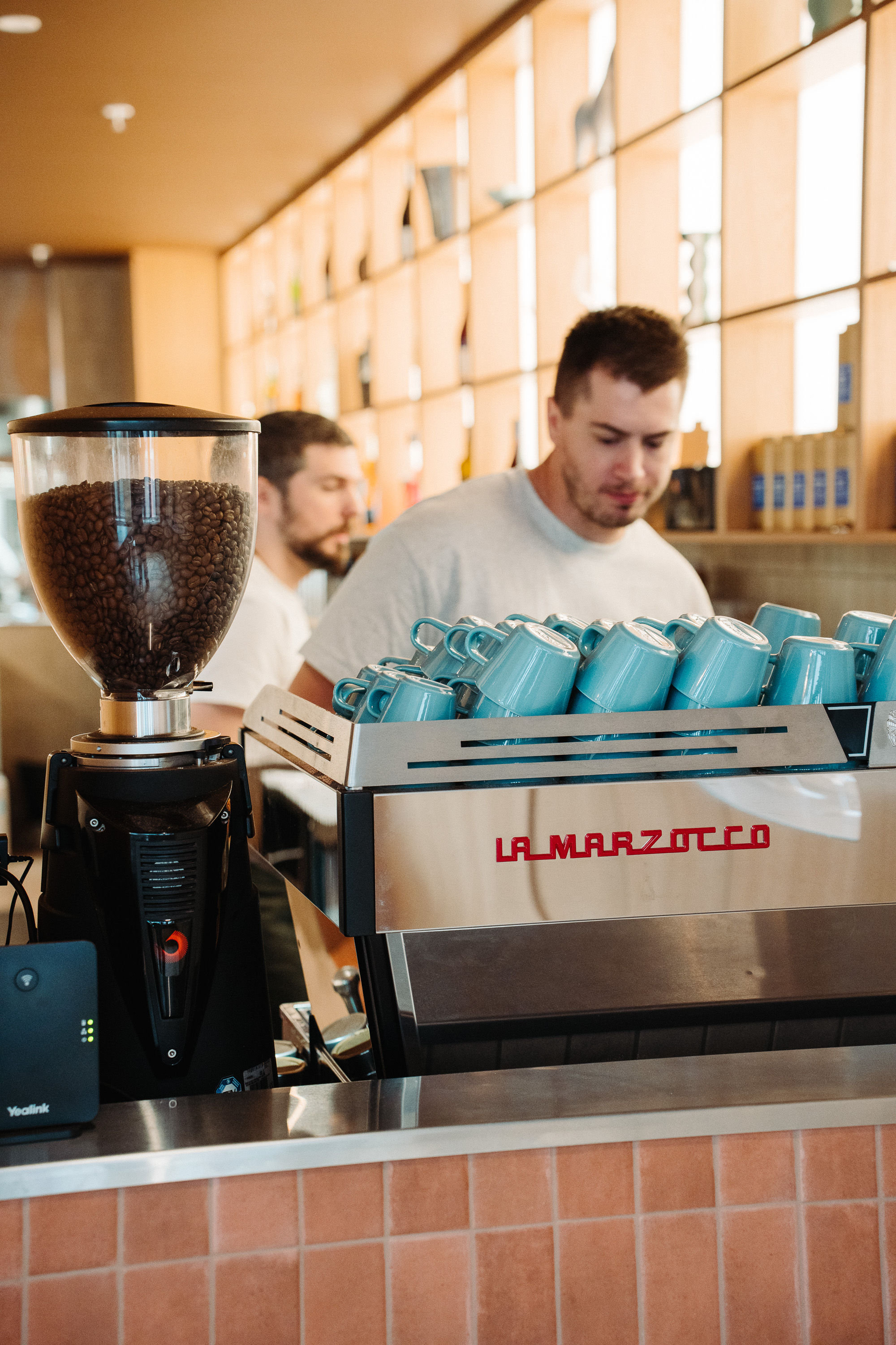A barista works behind an espresso machine with a row of blue cups on top, while a coffee grinder filled with beans sits in the foreground of a bright cafe.