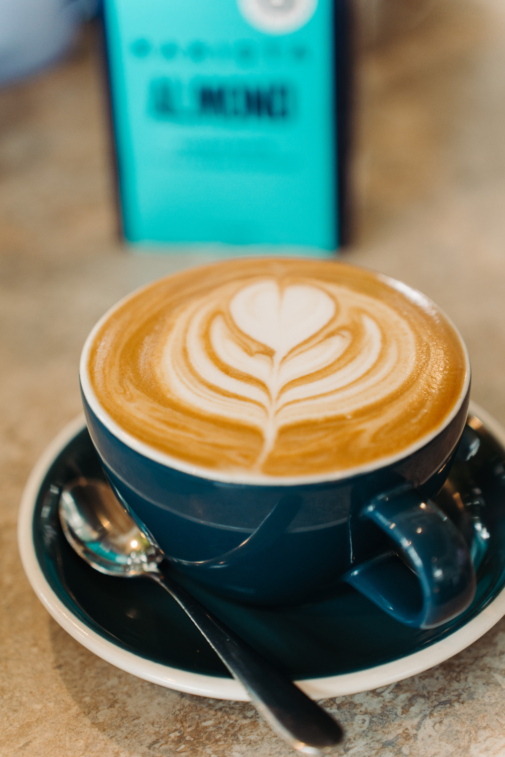 Close-up photo of a latte in a dark ceramic cup and saucer, featuring heart-shaped latte art on the foam; a teal menu sign is softly blurred in the background.