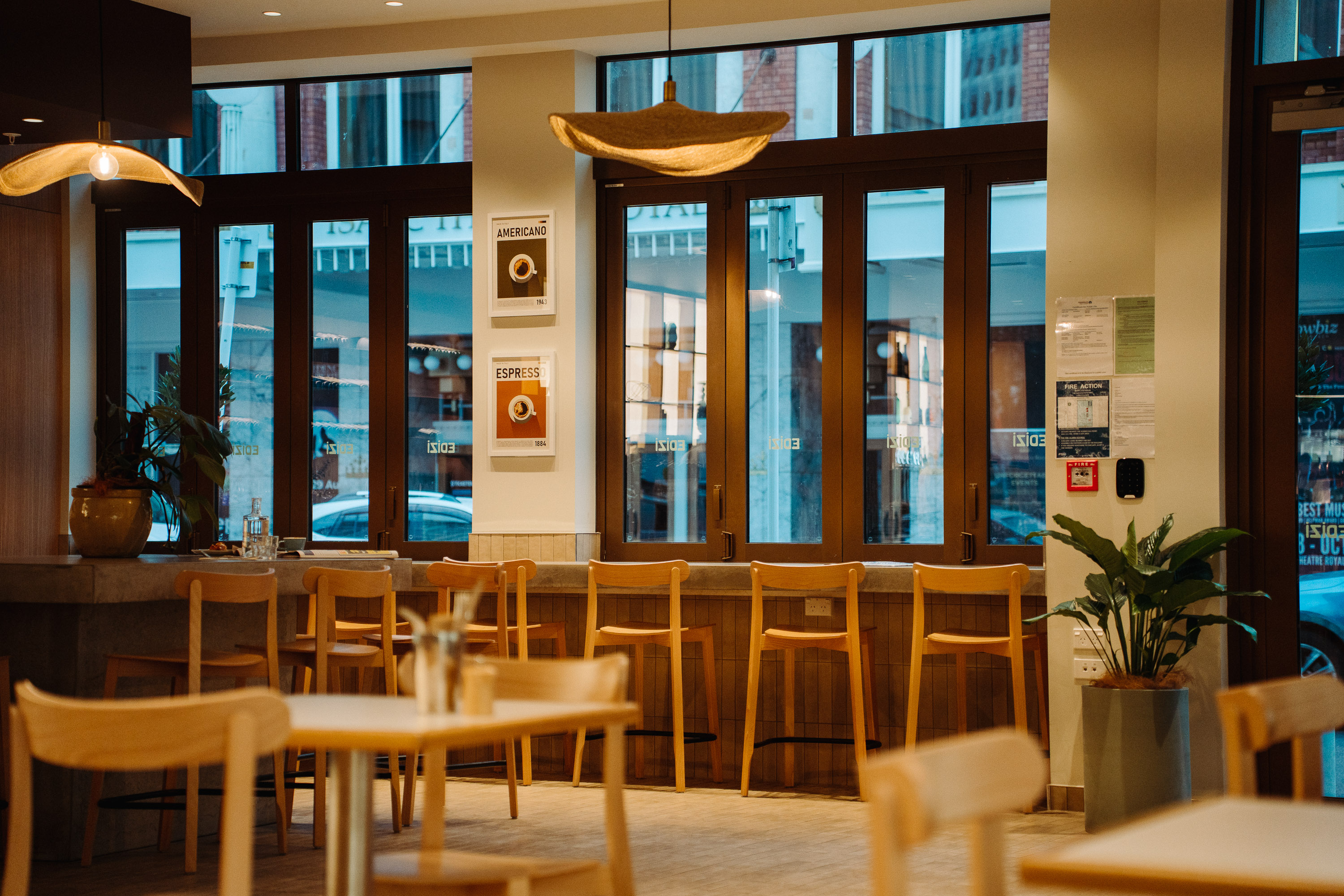 Interior of a modern cafe with wooden chairs and tables, warm pendant lighting, and large street-facing windows along a bar seating area.