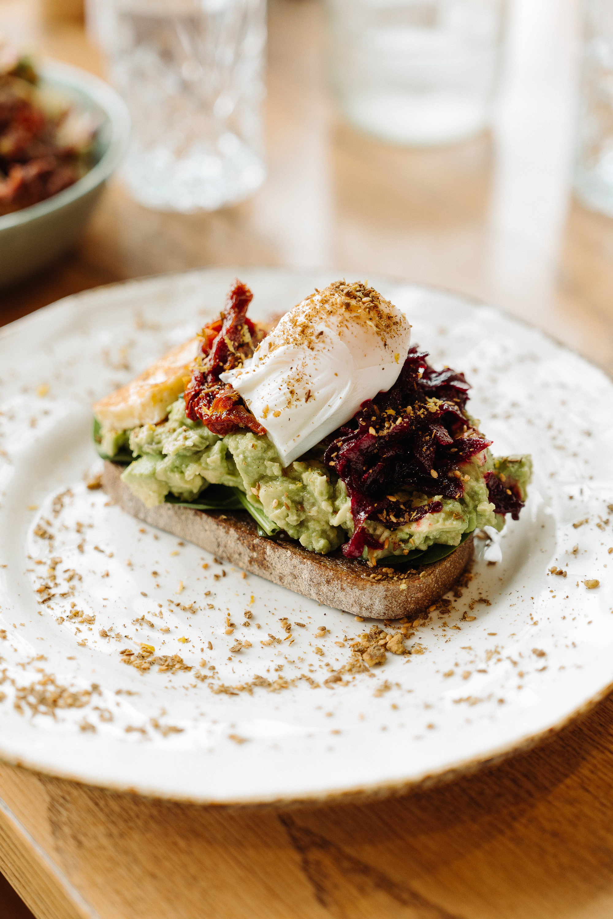 Close-up of avocado toast on a ceramic plate, topped with a poached egg, sun-dried tomatoes, and a shredded beet slaw, sprinkled with seasoning on a café table.