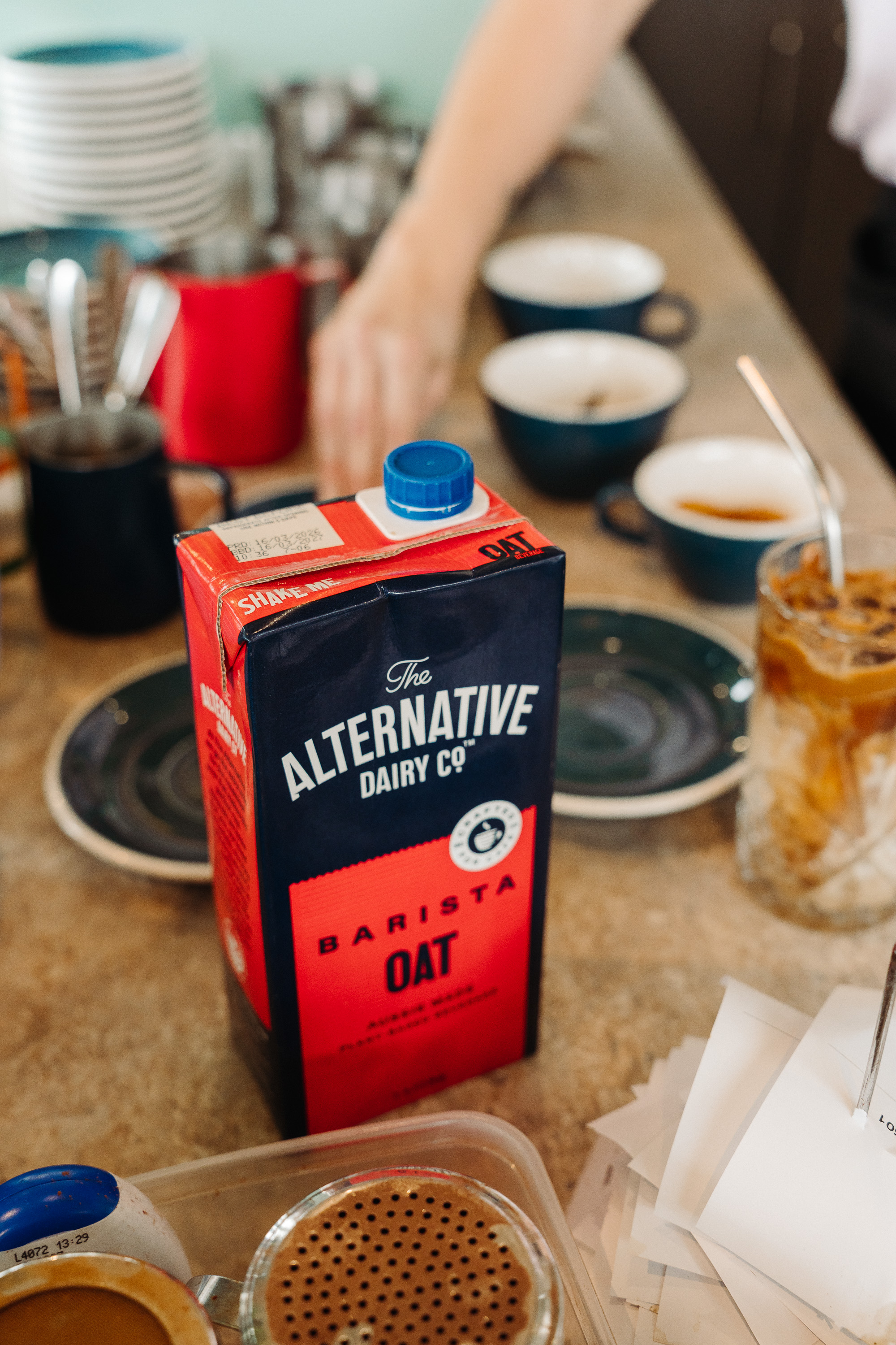 A carton of oat milk sits on a café counter beside cups and a glass of iced coffee, with a barista working in the background.