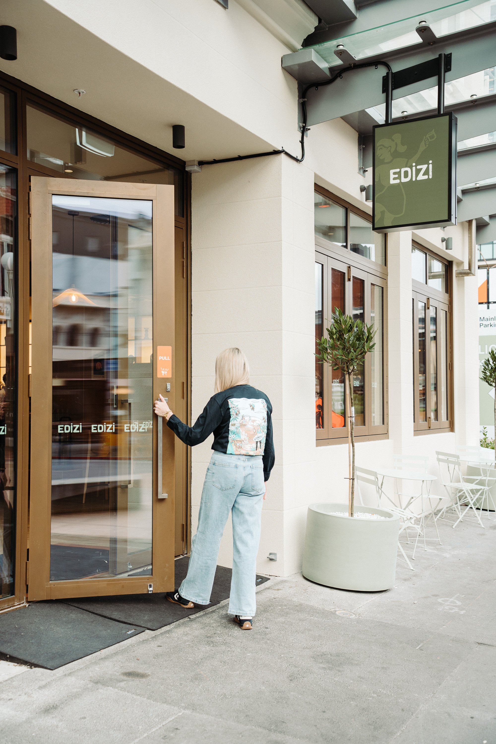 A person stands outside a modern storefront and reaches for the glass door handle beneath a hanging “EDIZI” sign. The scene shows a bright, clean walkway with large windows, a potted tree, and outdoor café-style seating nearby.