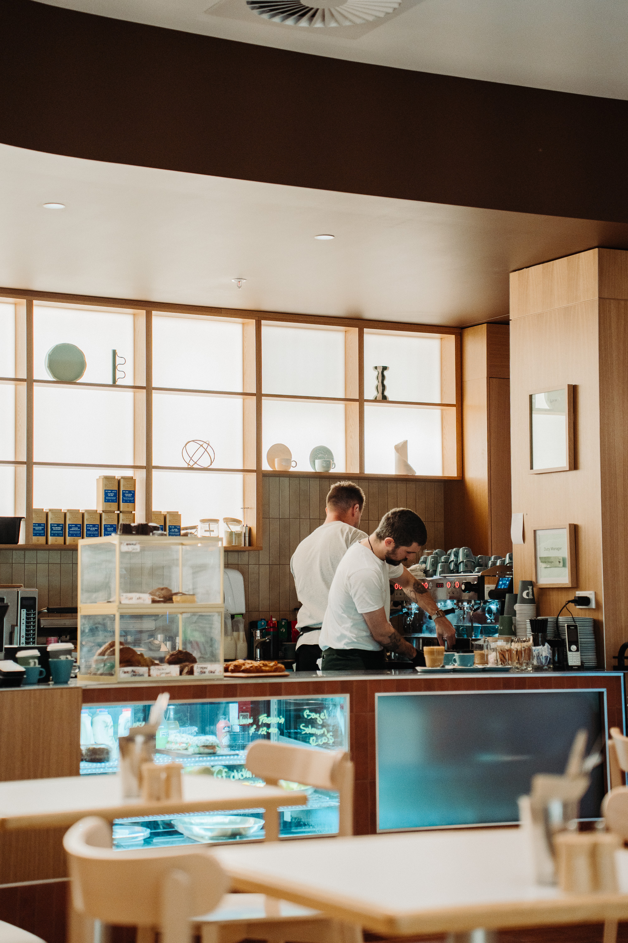 Warm, modern café interior with two baristas working behind the counter near an espresso machine. Pastries sit in a glass display case, and minimalist shelving and wood finishes create a cozy atmosphere.
