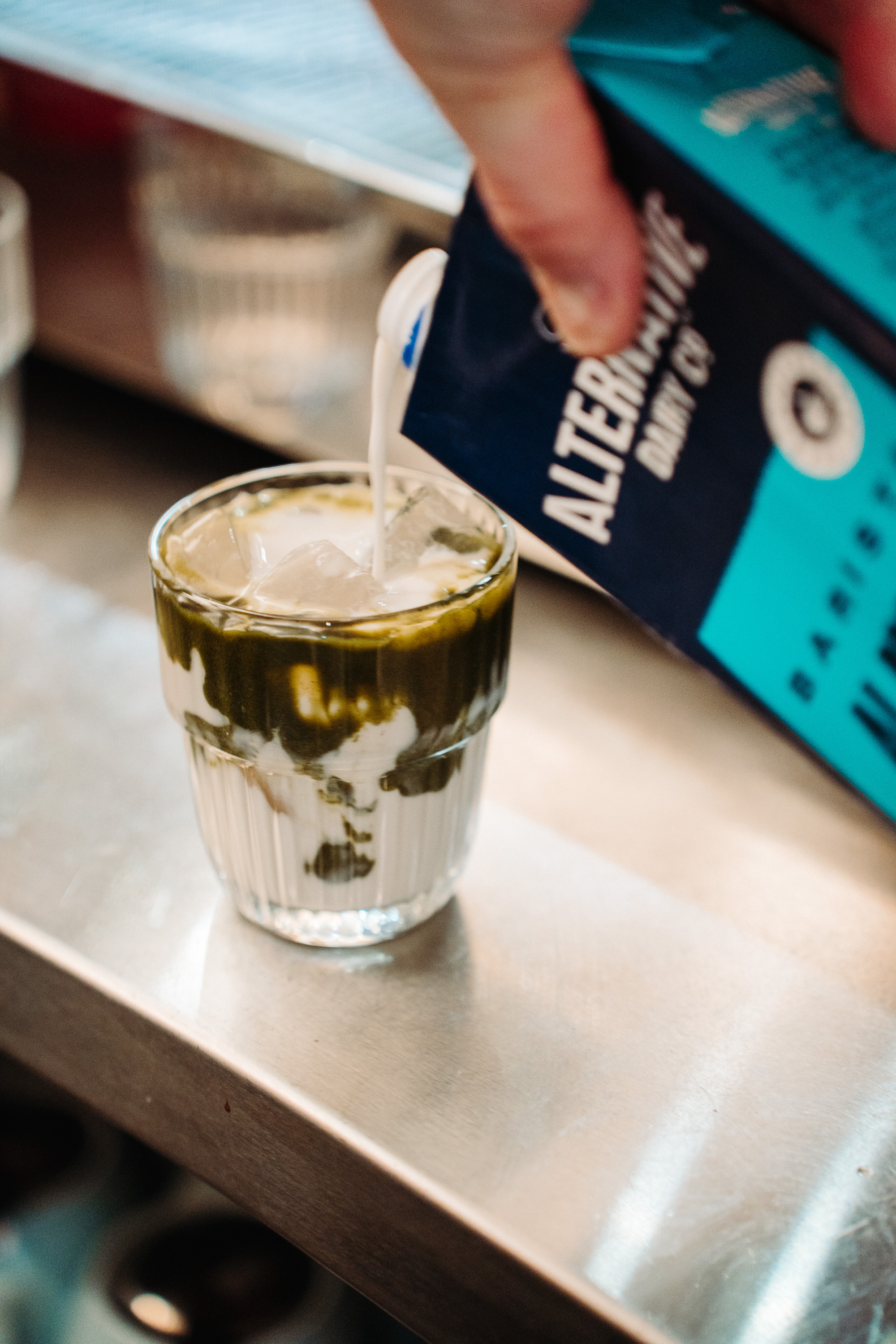 Close-up action shot of a hand pouring milk from a carton into a small glass filled with ice, creating a creamy drink with dark syrup or sauce swirled around the inside of the glass. The scene sits on a metal countertop, suggesting a café or drink-prep setting.