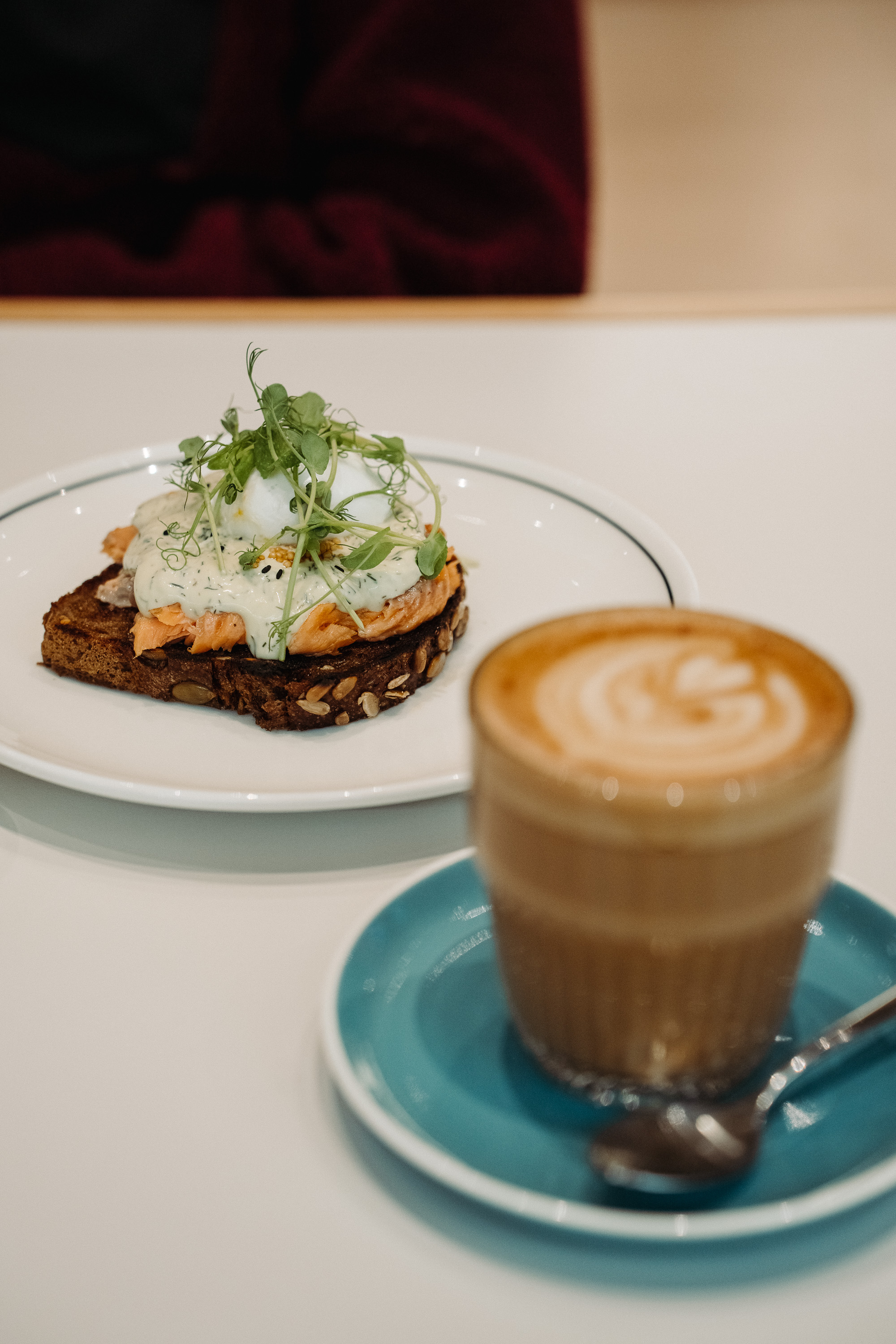 A plated open-faced sandwich on dark seeded bread topped with salmon, a creamy herb sauce, a poached egg, and microgreens, with a latte in the foreground on a blue saucer.