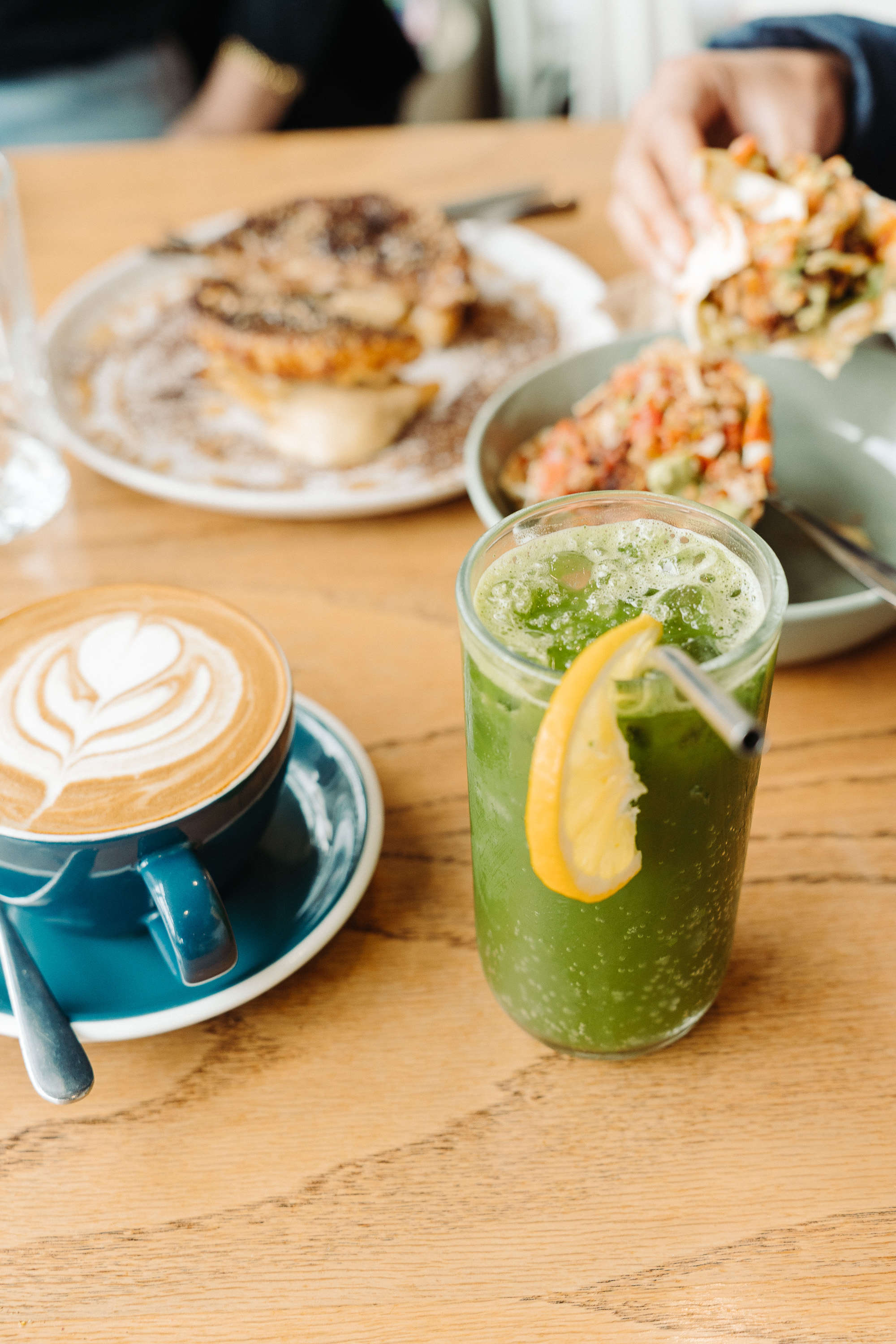 A café table with a cup of latte art and a tall iced green drink garnished with a lemon slice. In the background, plates of food and a person’s hands are softly out of focus.