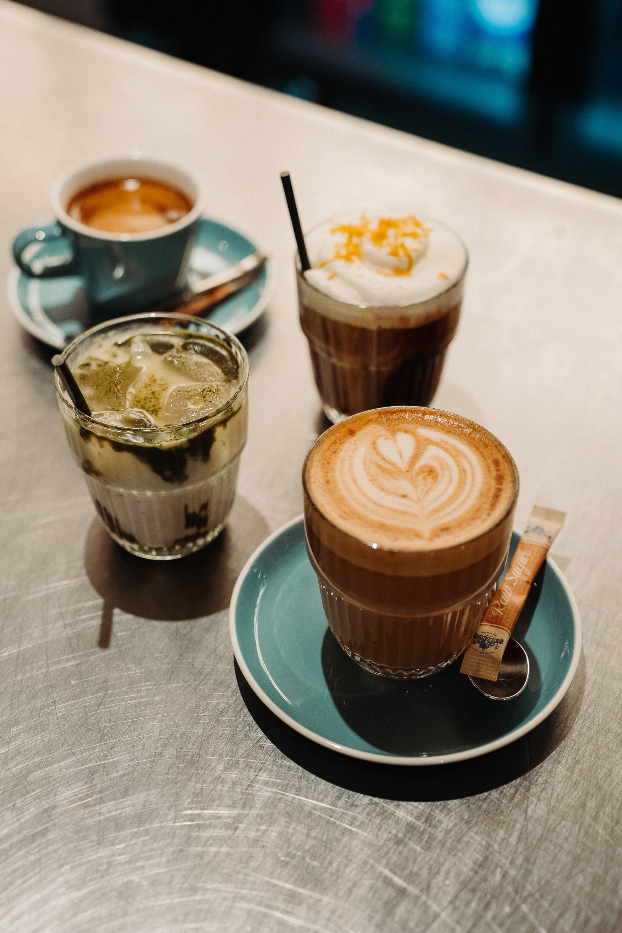 A close-up view of four café drinks on a metallic table: a latte with heart latte art, an iced matcha-style drink, a small espresso, and a layered iced coffee topped with whipped cream and citrus zest.