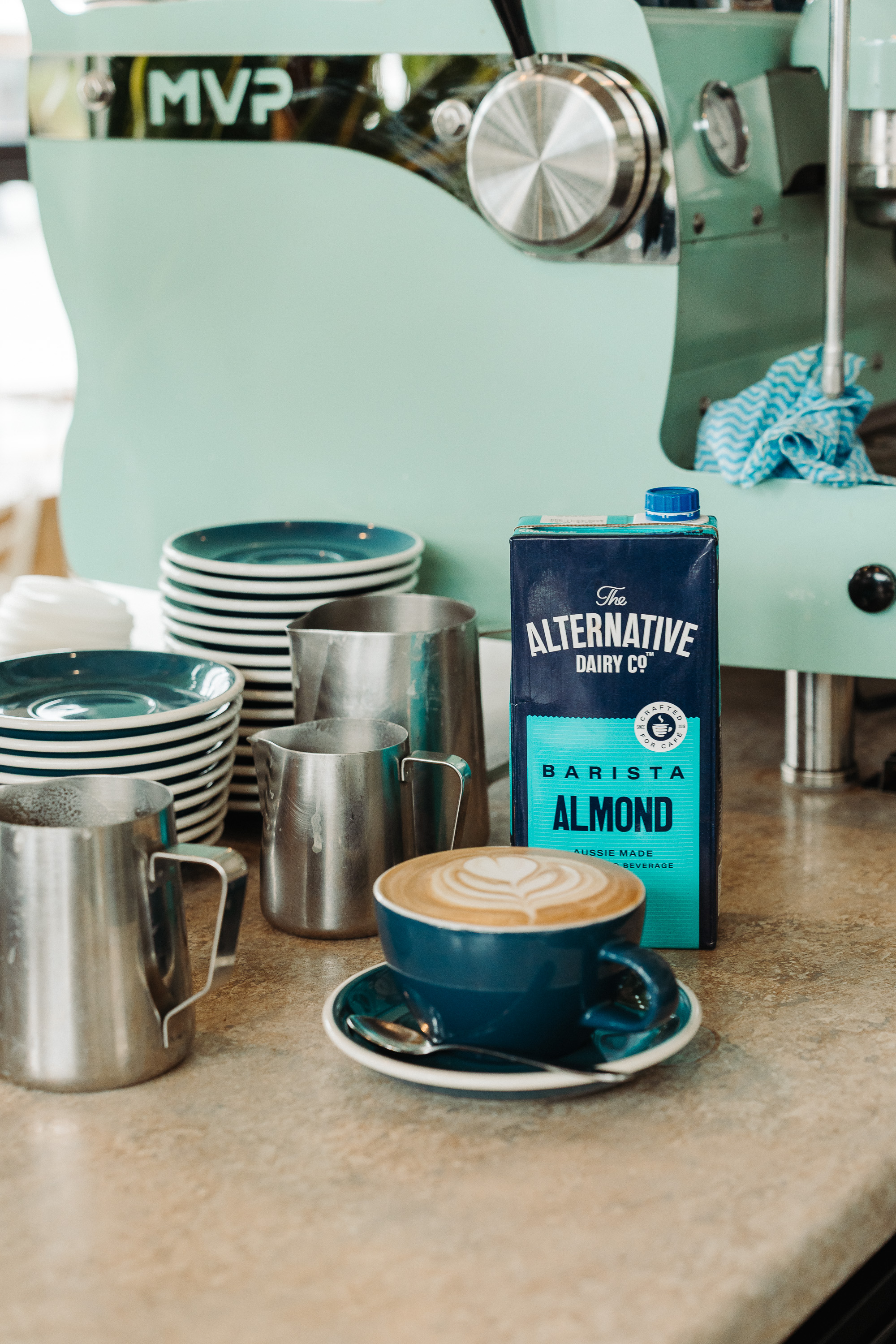 Close-up of a café counter with a mint-green espresso machine, stacked plates, stainless-steel milk pitchers, a latte with heart-shaped latte art, and a carton of almond milk.