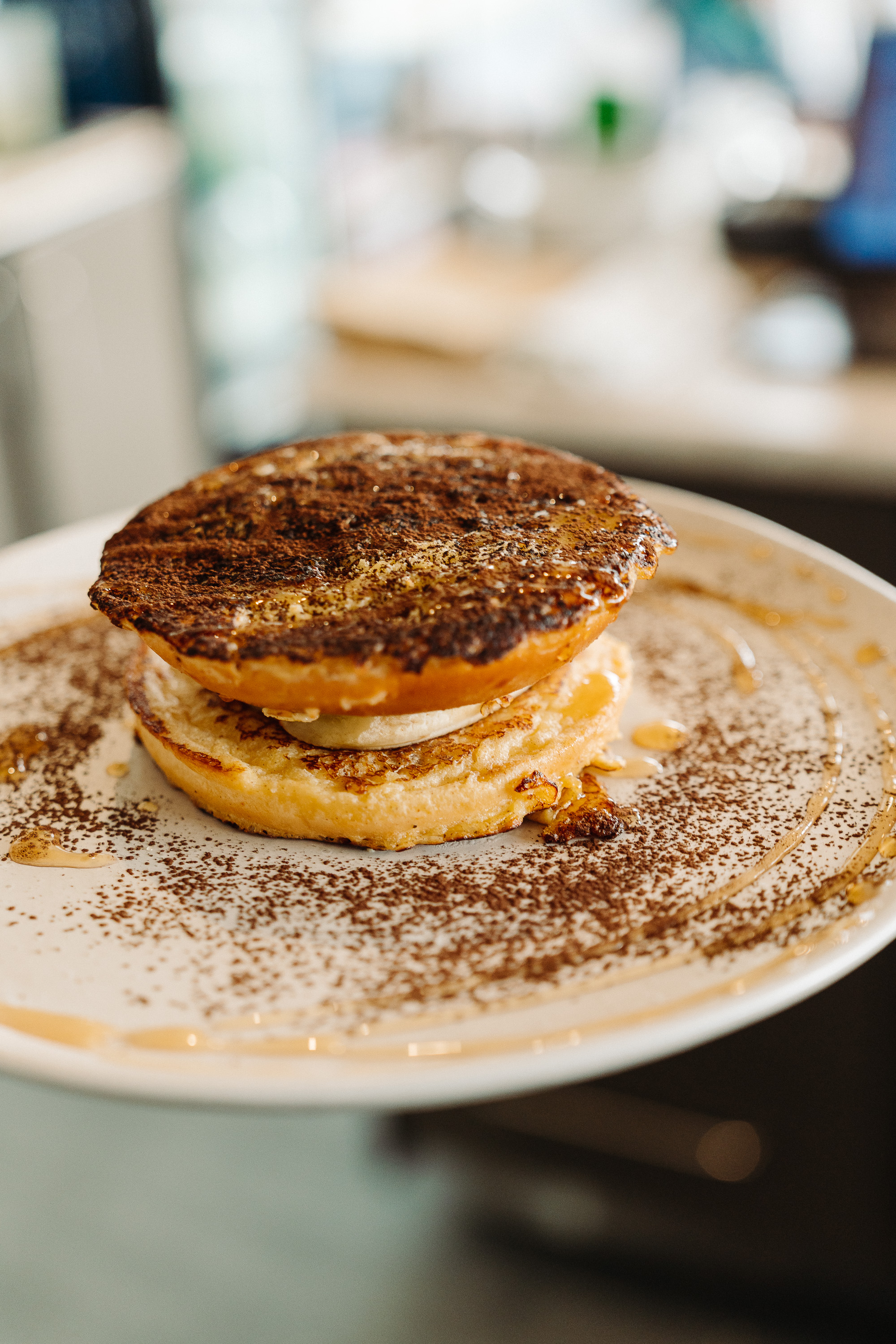 Close-up of a stacked breakfast dish on a white plate, dusted with cocoa or cinnamon and drizzled with syrup, with a softly blurred café background.