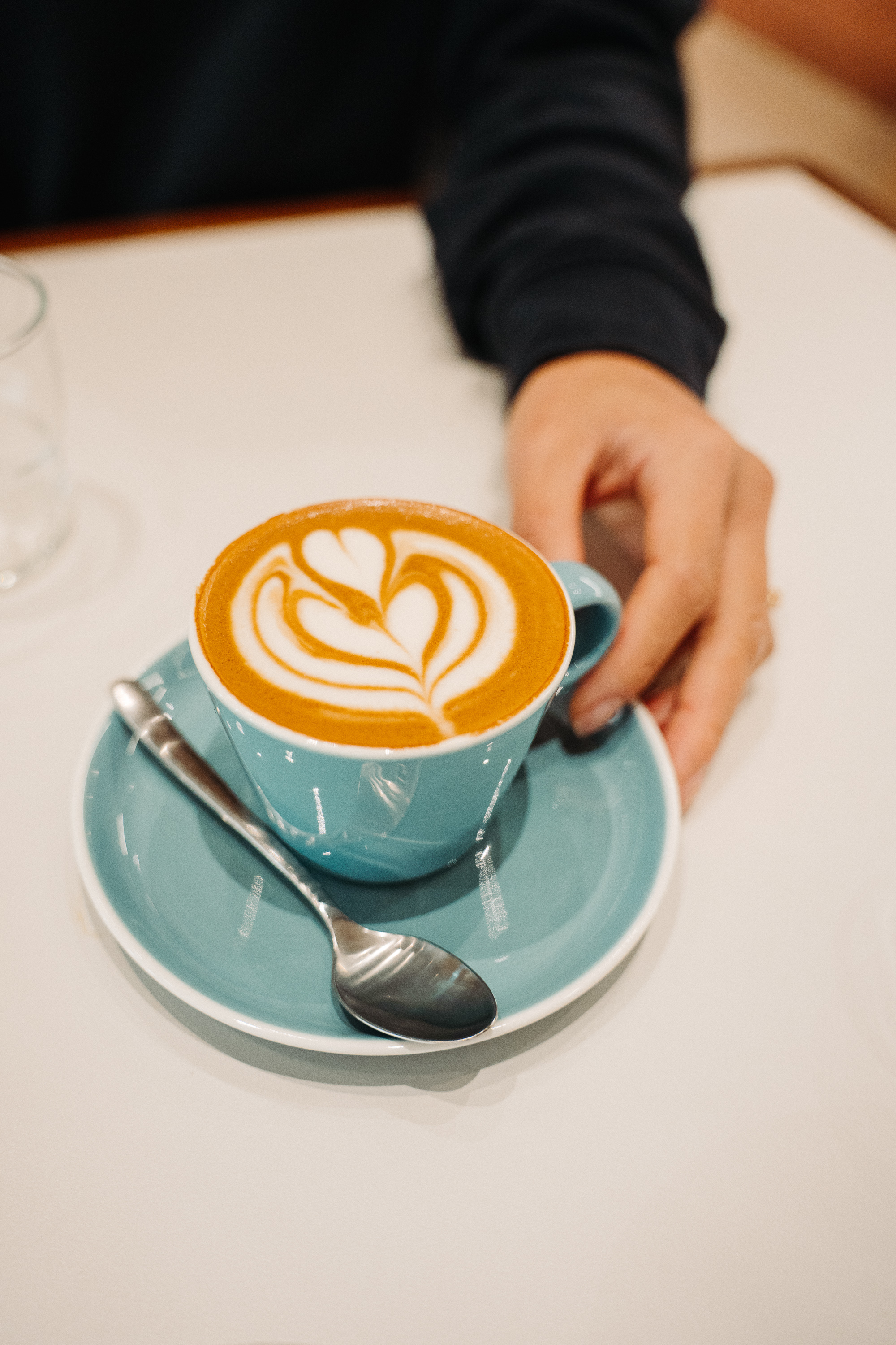A hand slides a teal ceramic cup and saucer across a café table, showing a latte topped with heart-shaped latte art and a teaspoon resting on the saucer.
