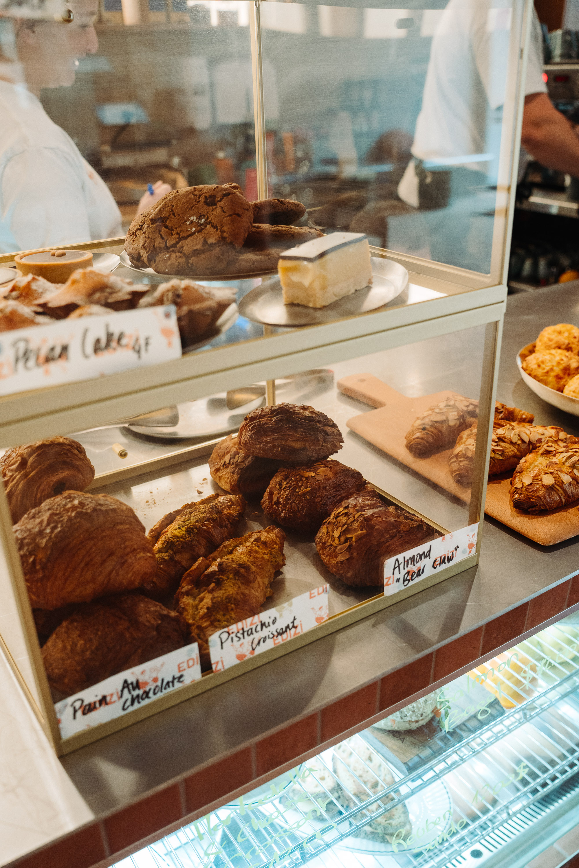 A glass display case filled with assorted pastries and baked goods, including croissants and other flaky pastries, with handwritten flavor labels visible on the trays. Blurred staff members stand behind the counter, giving the scene a warm, busy bakery or café atmosphere.
