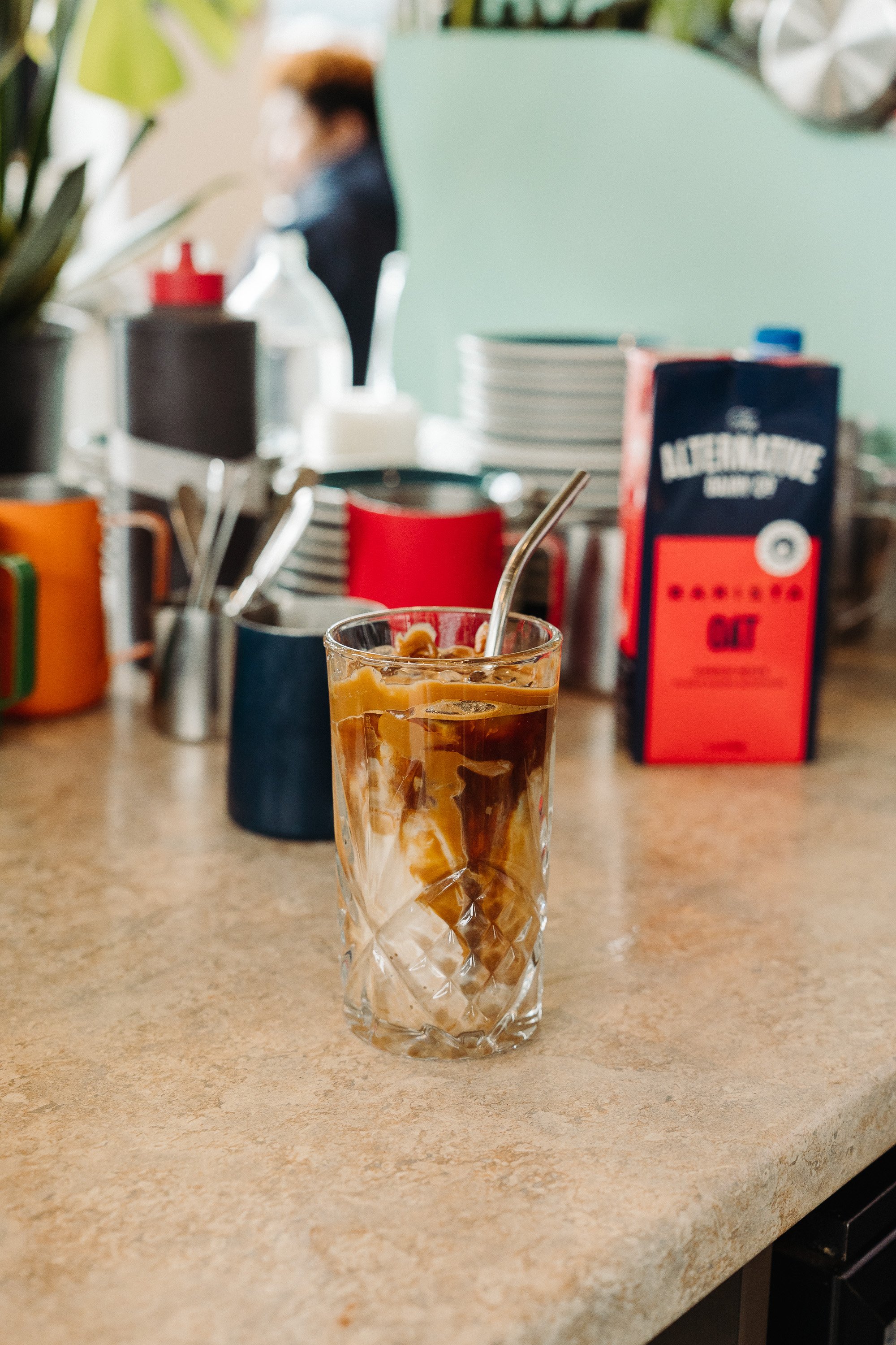 A tall glass of iced coffee with milk swirling through it and a metal straw, sitting on a café counter with softly blurred mugs, utensils, and coffee packaging in the background.