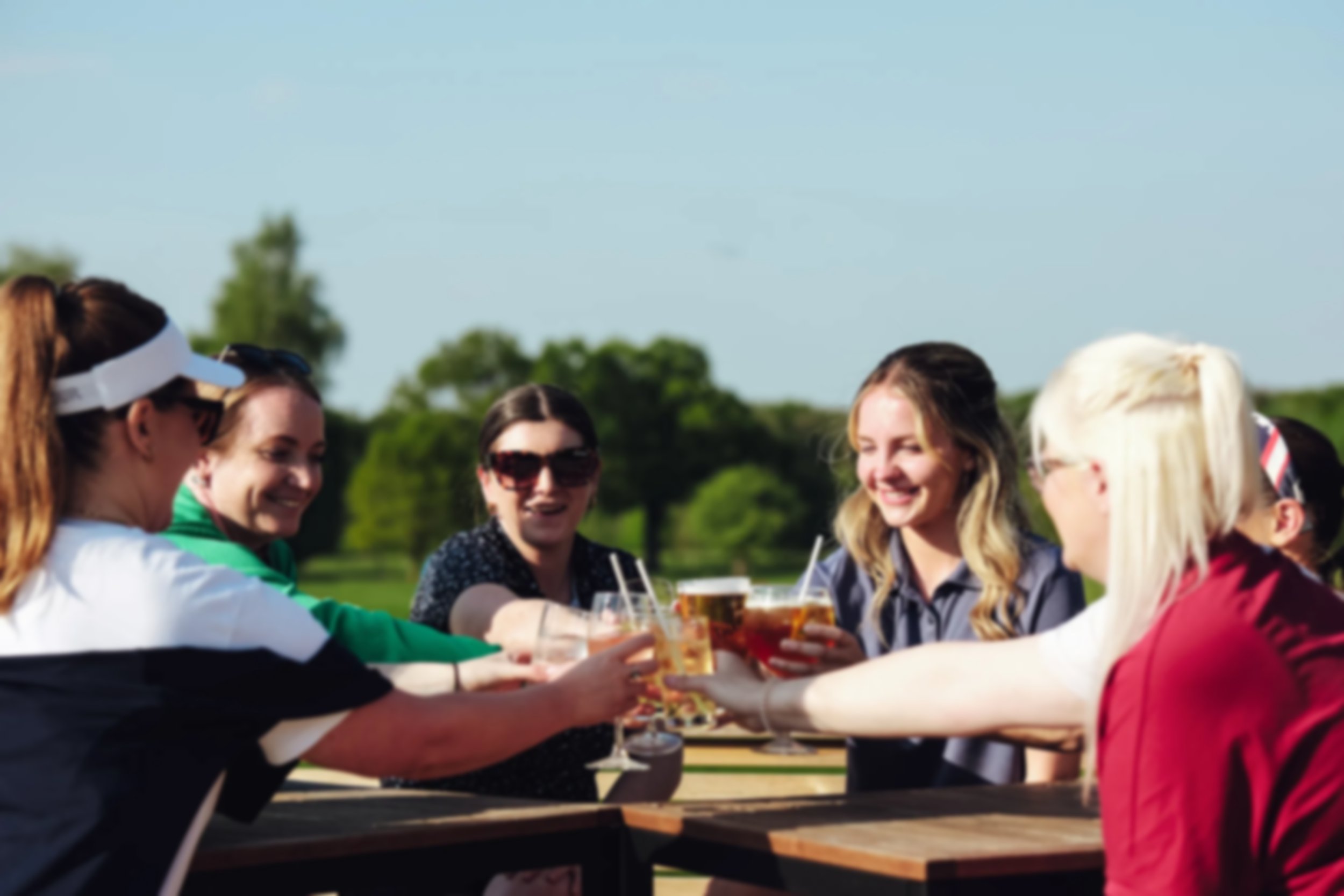 Women golfers sharing drinks and laughs on a sunny day with lush greenery in the background.