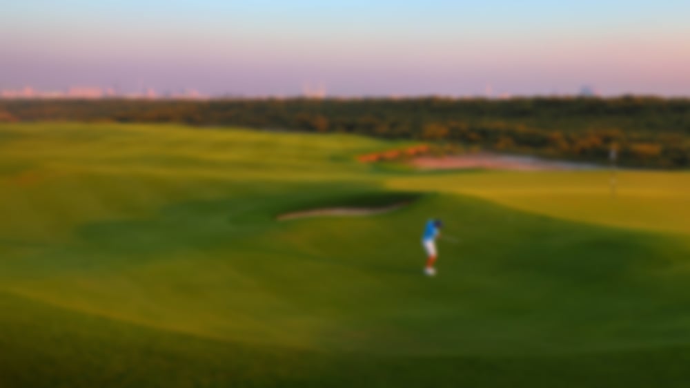 Golfer chipping onto the elevated green with the UAE backdrop and sunset.