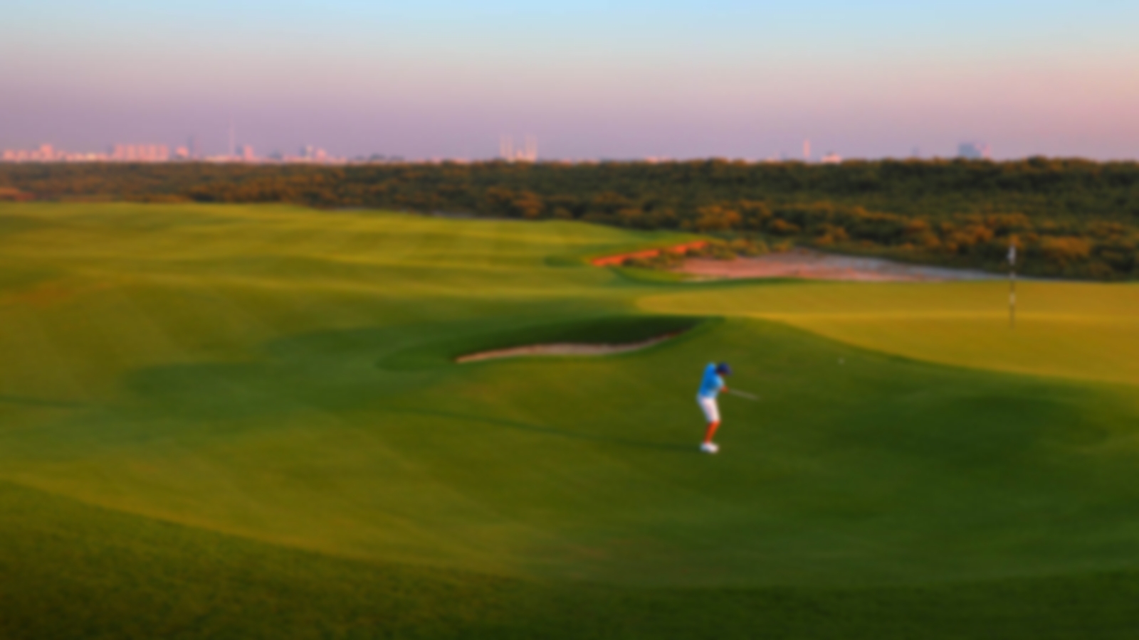 Golfer chipping onto the elevated green with the UAE backdrop and sunset.