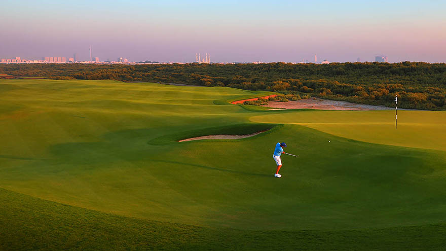 Golfer chipping onto the elevated green with the UAE backdrop and sunset.