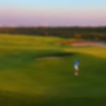 Golfer chipping onto the elevated green with the UAE backdrop and sunset.