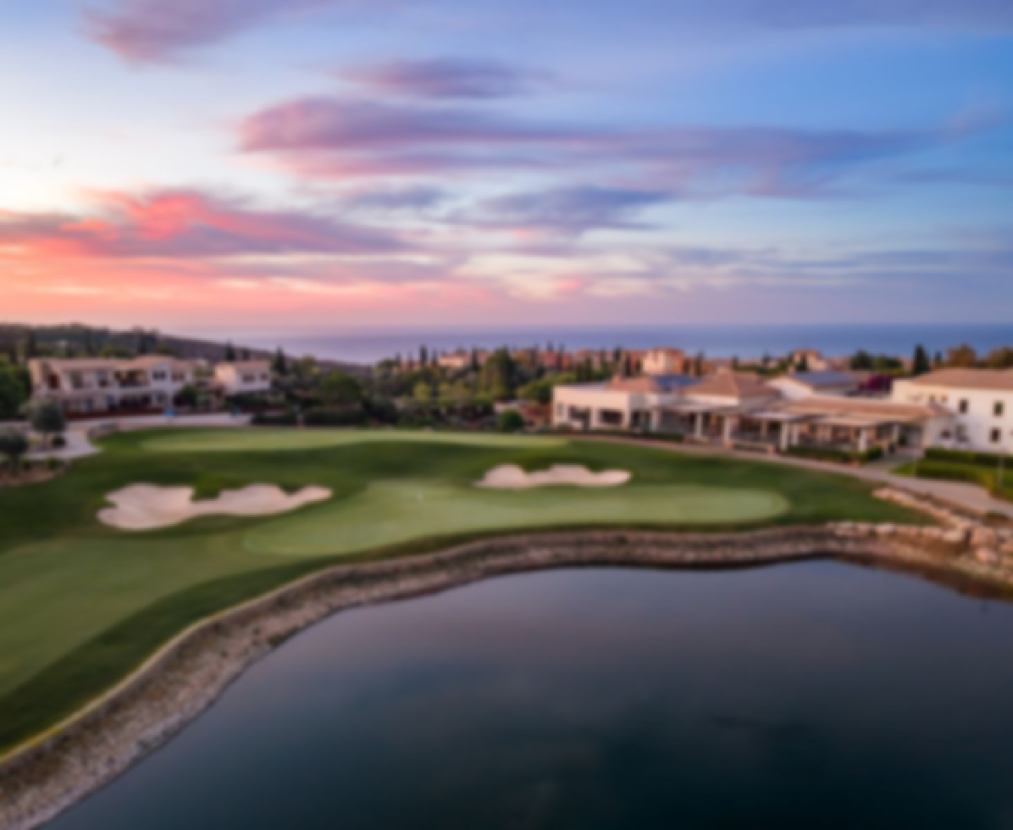The 18th green at Aphrodite Hills beside a water feature and clubhouse, with a colourful sunset over the sea.