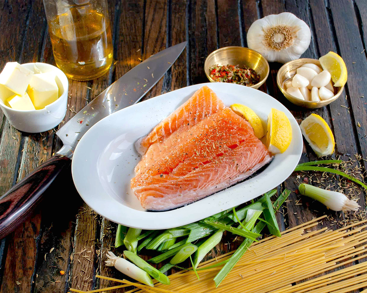 Food display of raw salmon filets and lemons on plate, garlic, cubed butter, seasonings, and various ingredients on a table.