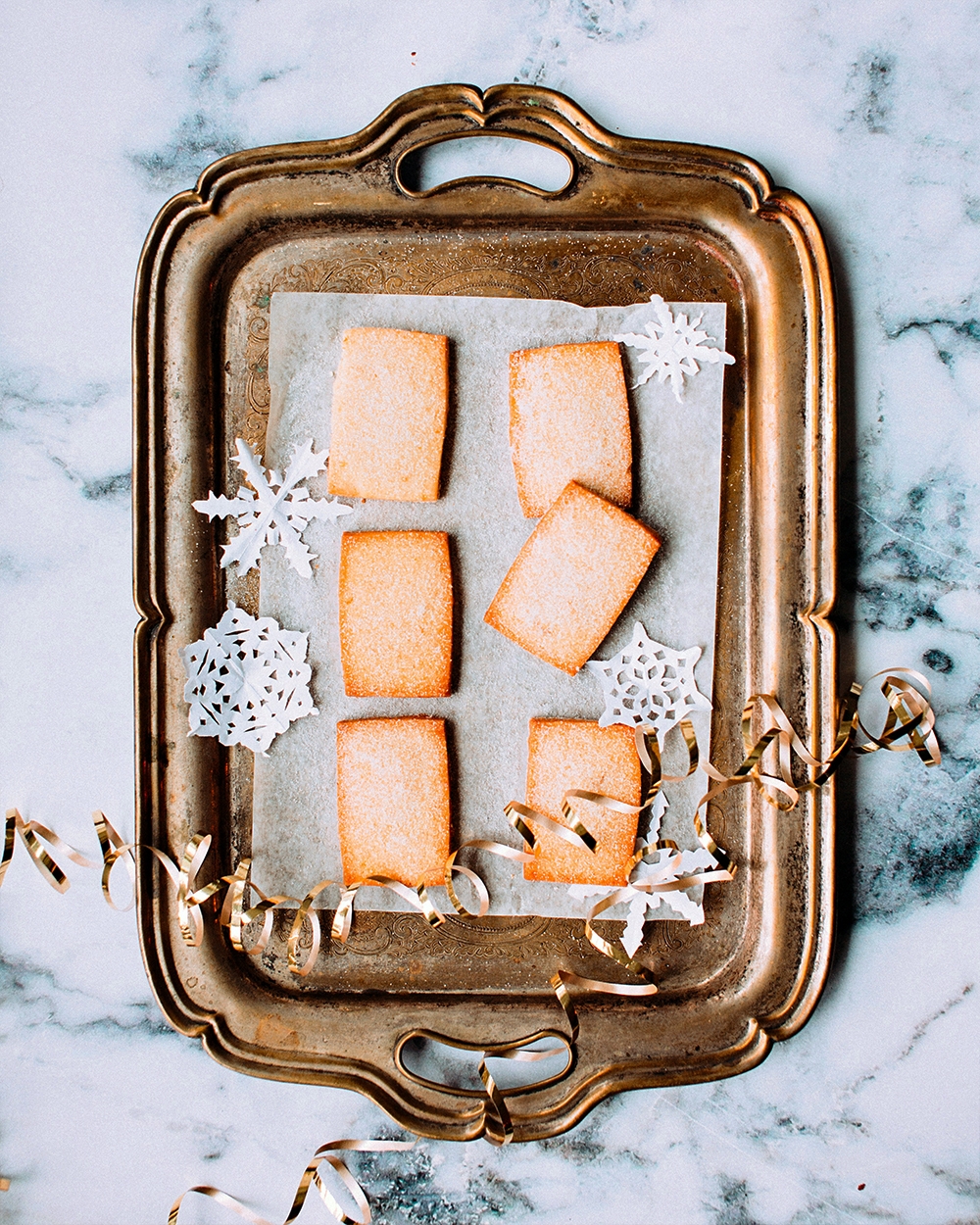 Bronze serving tray with parchment paper, rectangle cookies, gold ribbon, and white paper snowflakes.