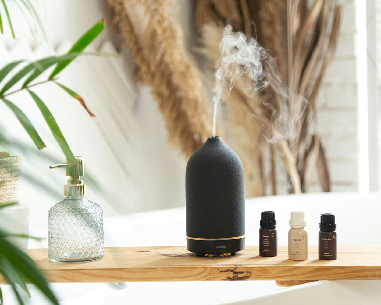 Wooden shelf displaying a black aromatherapy diffuser, small bottles of essential oils, and glass soap dispenser.  
