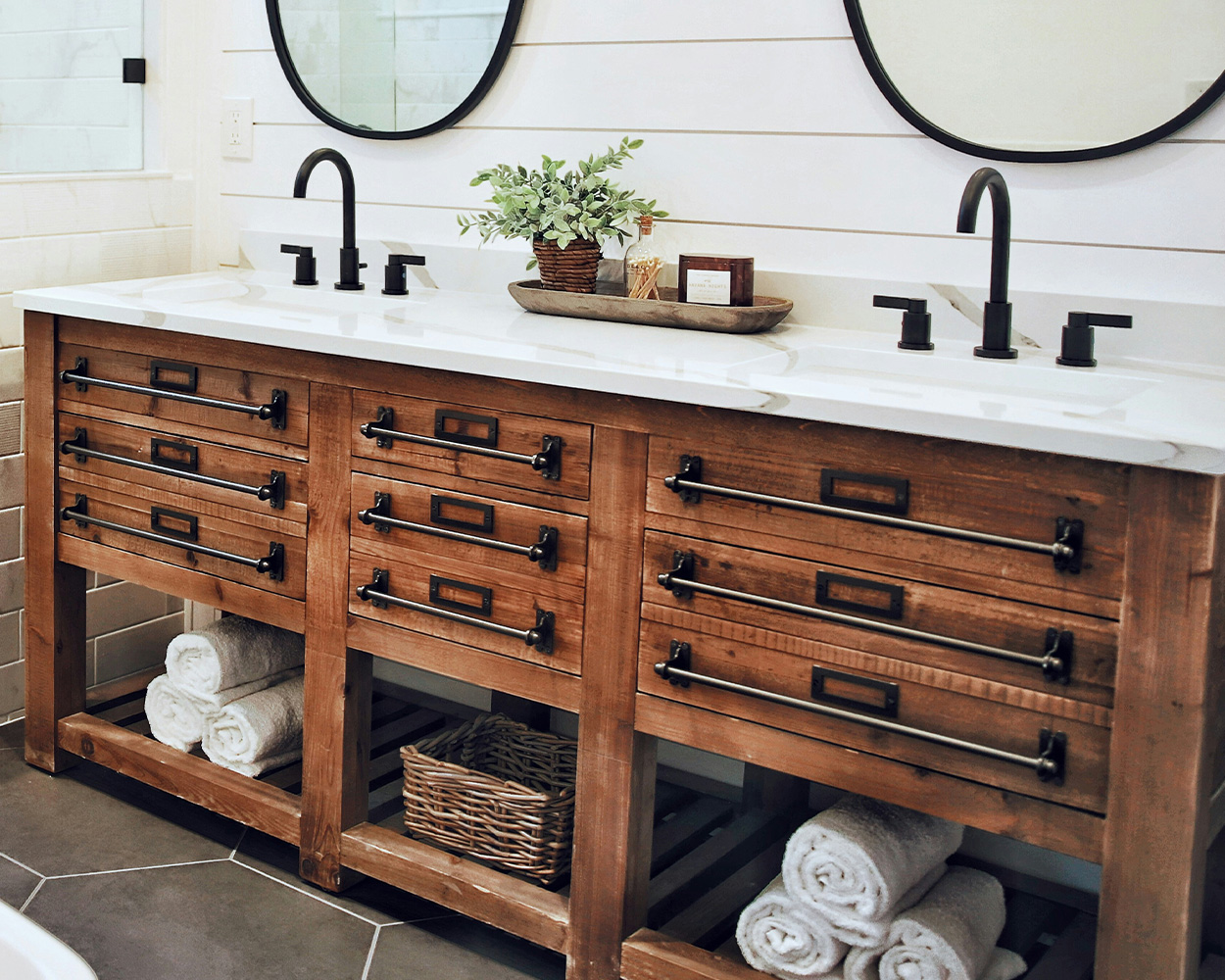 Wooden bathroom vanity with marble countertop, two sinks, nine drawers, organized towels, wicker basket, and decor.