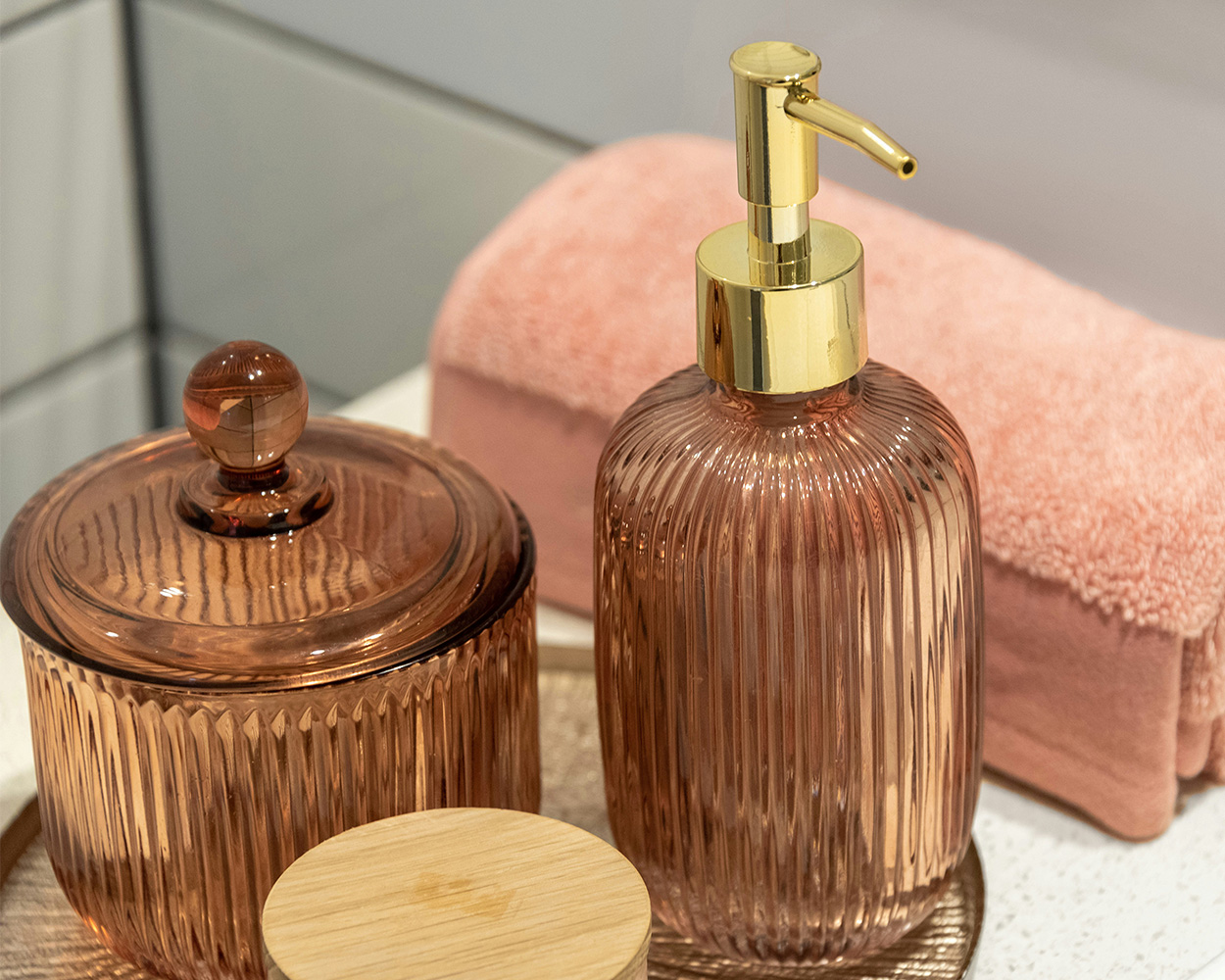 Dark pink glass soap dispenser and container with lid sitting on a bathroom counter next to a light pink rolled towel. 
